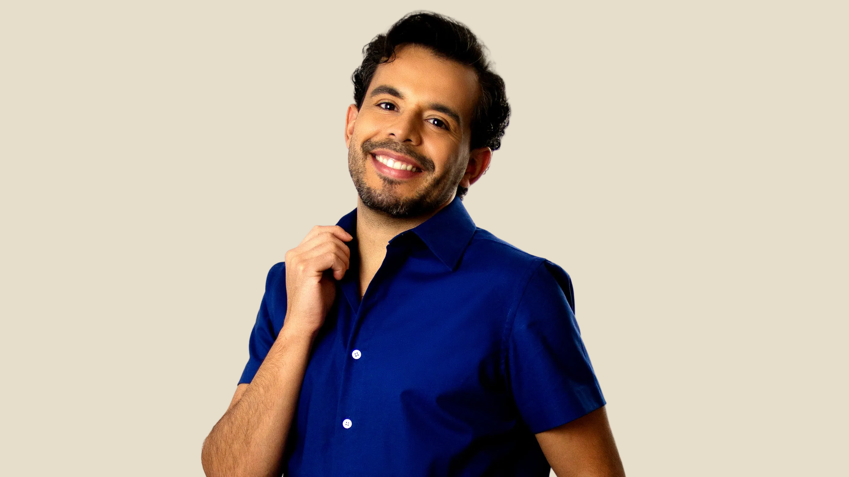 A smiling man with dark hair and a beard wearing a royal blue short-sleeve button-up shirt, posing against a solid beige background.